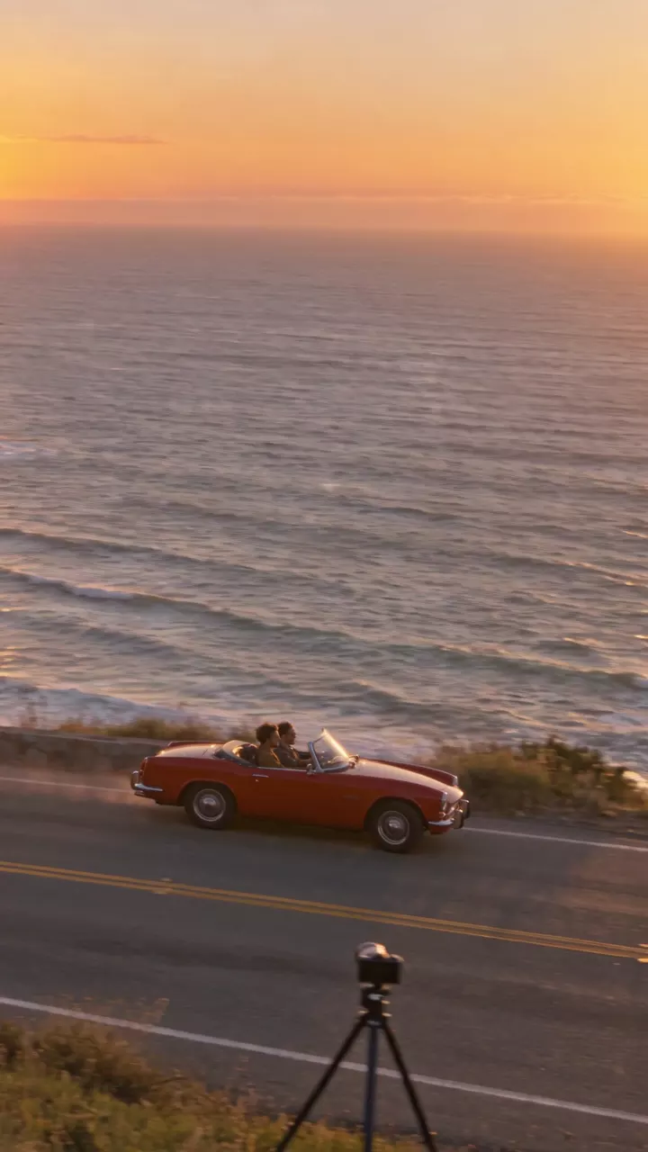 Take 2, the coast road at sunset: the camera turns with the red convertible, and a camera tripod appears standing by the road